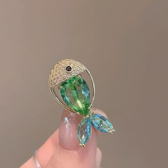 A close-up of a hand holding a fish-shaped brooch with a gold-tone setting, a green faceted crystal body, and light blue crystal tail fins against a plain taupe background.