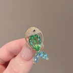 A hand holding a gold fish-shaped brooch with a green teardrop crystal body, a gold crystal-encrusted head, and two light blue crystal fins, against a plain taupe background.