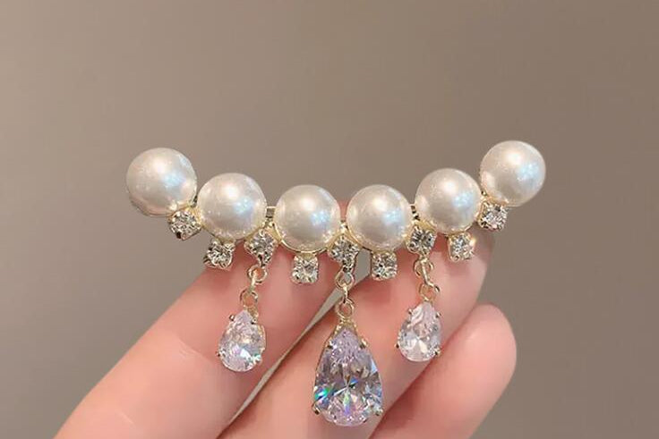 A close-up of a pearl and crystal hair claw clip being held on a person's fingers against a soft beige background. The clip features five white pearls, small clear crystals, and three dangling teardrop-shaped clear crystals.