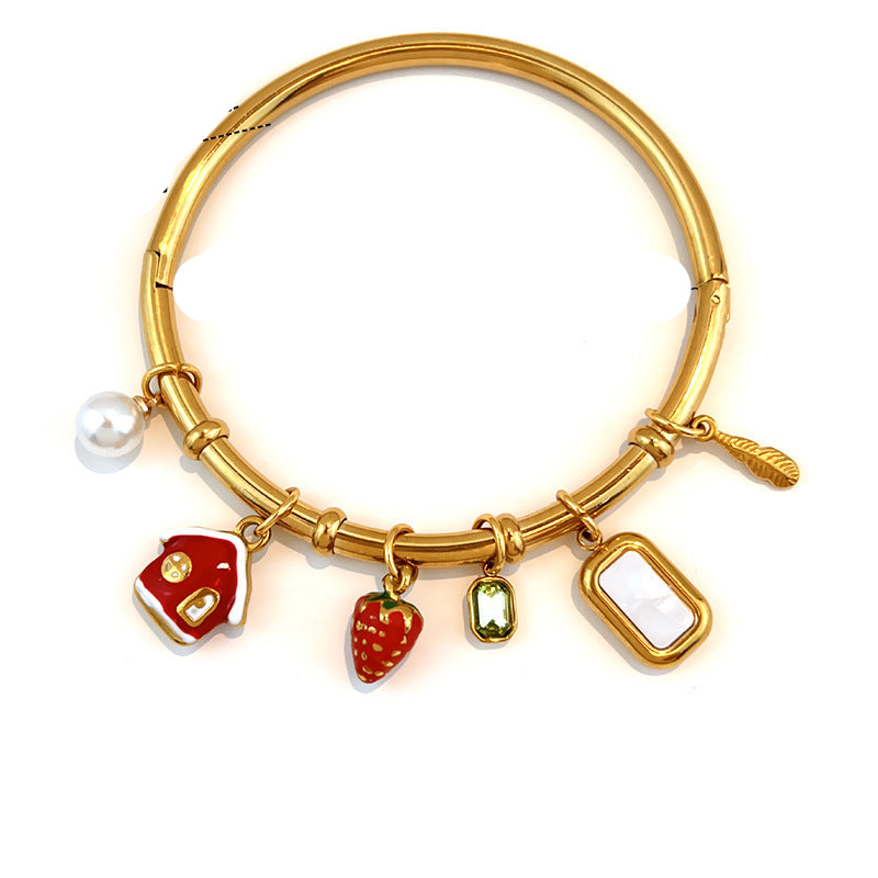 Gold-plated stainless steel bangle bracelet with four enamel charms (a red house, a strawberry, a green teardrop, and a white rectangular charm) and a pearl bead, shown with dimension markings of 6.0cm width and 0.
